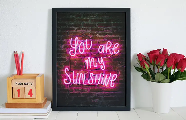 Cadre noir encadrant une néon rose "You are my sunshine" sur mur brique, cadeau personnalisé idéal pour la Saint-Valentin.
