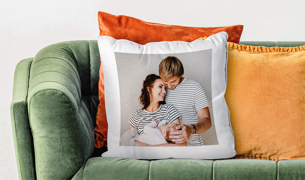 Oreiller personnalisé avec photo de famille : un couple heureux câlinant leur bébé, imprimé sur un coussin blanc posé sur un canapé vert.